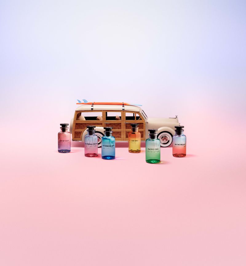 A collection of six colorful Louis Vuitton glass perfume bottles arranged before a miniature wooden woody station wagon with a surfboard.