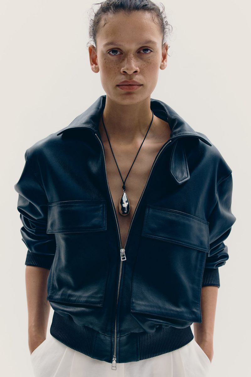 Model with pulled-back hair wearing a black leather zip-up jacket and a silver pendant on a cord against a plain white background.