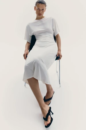 Model Mila van der Horst seated on a chair wearing a white short-sleeve dress and black heeled sandals against a white background.