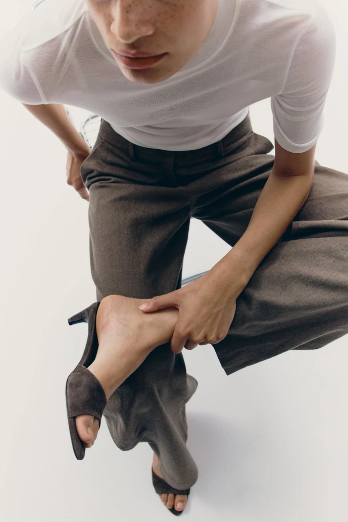 High-angle view of model Mila van der Horst in brown trousers and suede mule heels against a white studio background.
