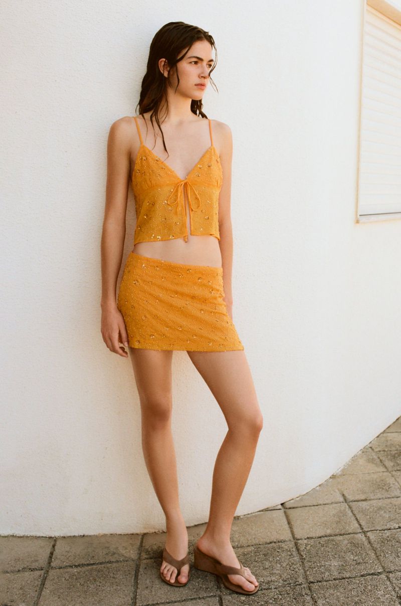 A woman stands against a white curved wall wearing a yellow floral camisole top and matching mini skirt with brown thong sandals.