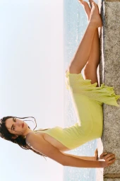 Model reclining in a sleeveless light yellow dress along a stone ledge overlooking sparkling ocean water.
