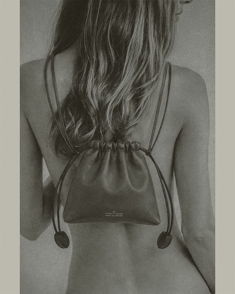 Model with long hair seen from behind wearing a dark leather drawstring mini backpack against a neutral studio backdrop.