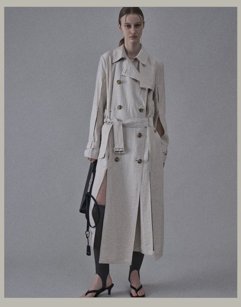 Model Maria Shainer poses in a light beige deconstructed belted trench coat and black heels against a grey studio backdrop.