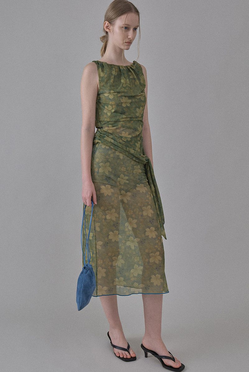 Model wearing a green floral semi-sheer sleeveless midi dress and black kitten heels against a neutral grey studio backdrop.