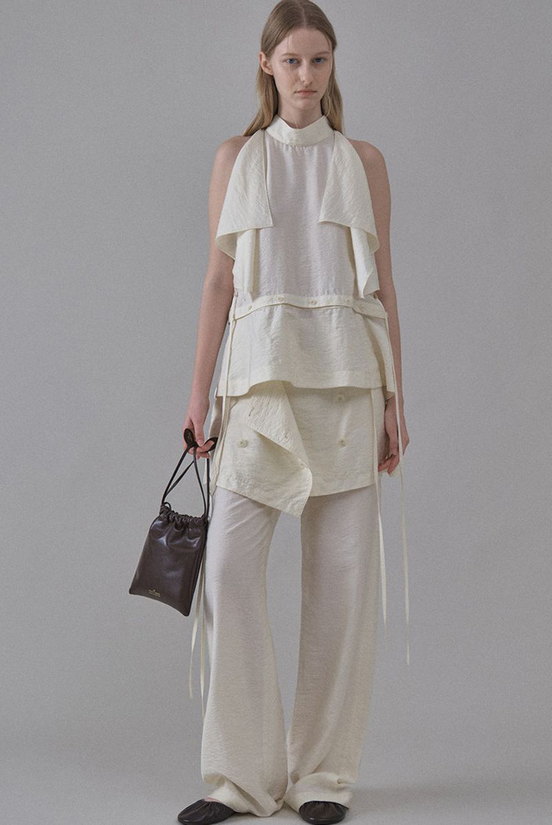 Model Maria Shainer poses in a cream layered sleeveless top and wide-leg trousers holding a dark brown pouch bag.