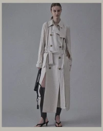 Model Maria Shainer poses in a light beige deconstructed belted trench coat and black heels against a grey studio backdrop.