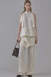 Model Maria Shainer poses in a cream layered sleeveless top and wide-leg trousers holding a dark brown pouch bag.