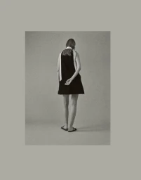 Model Maria Shainer seen from behind wearing a short black dress with white scarf detail and flat sandals against a neutral studio backdrop.