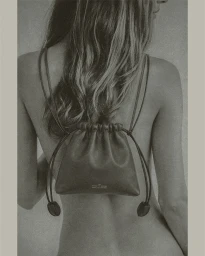 Model with long hair seen from behind wearing a dark leather drawstring mini backpack against a neutral studio backdrop.