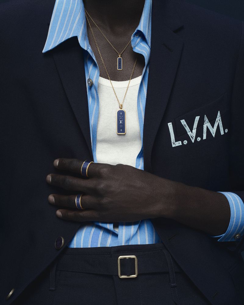 A Black male model wears a navy blazer with L.V.M. embroidery, a blue striped shirt, white tank top, and Louis Vuitton gold jewellery.