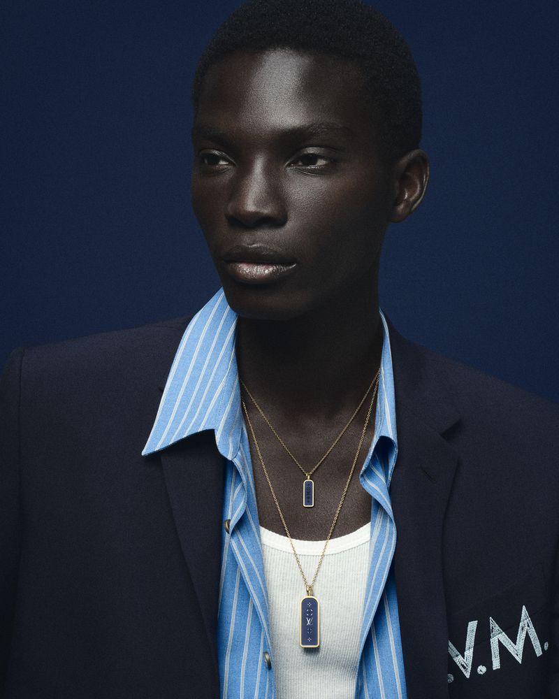 Model Dara Gueye wearing layered gold pendant necklaces, a blue striped shirt, and a navy blazer against a dark blue backdrop.
