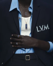 Close-up of a model in a navy blazer and blue striped shirt wearing Louis Vuitton gold and blue pendant necklaces and matching rings.