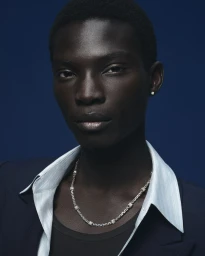 Dara Gueye wearing a silver chain necklace, open striped shirt, and navy blazer against a deep blue background for Louis Vuitton.