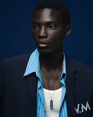 Model Dara Gueye wearing layered gold pendant necklaces, a blue striped shirt, and a navy blazer against a dark blue backdrop.