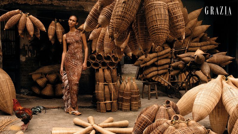 A striking fashion editorial shot outdoors featuring a model in a patterned dress, surrounded by traditional woven fishing baskets.