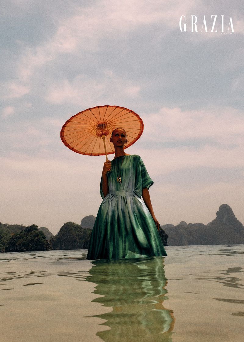 A lone fashion model stands in ocean water holding a parasol in front of a mountainous coast under a cloudy sky. Editorial fashion shot.