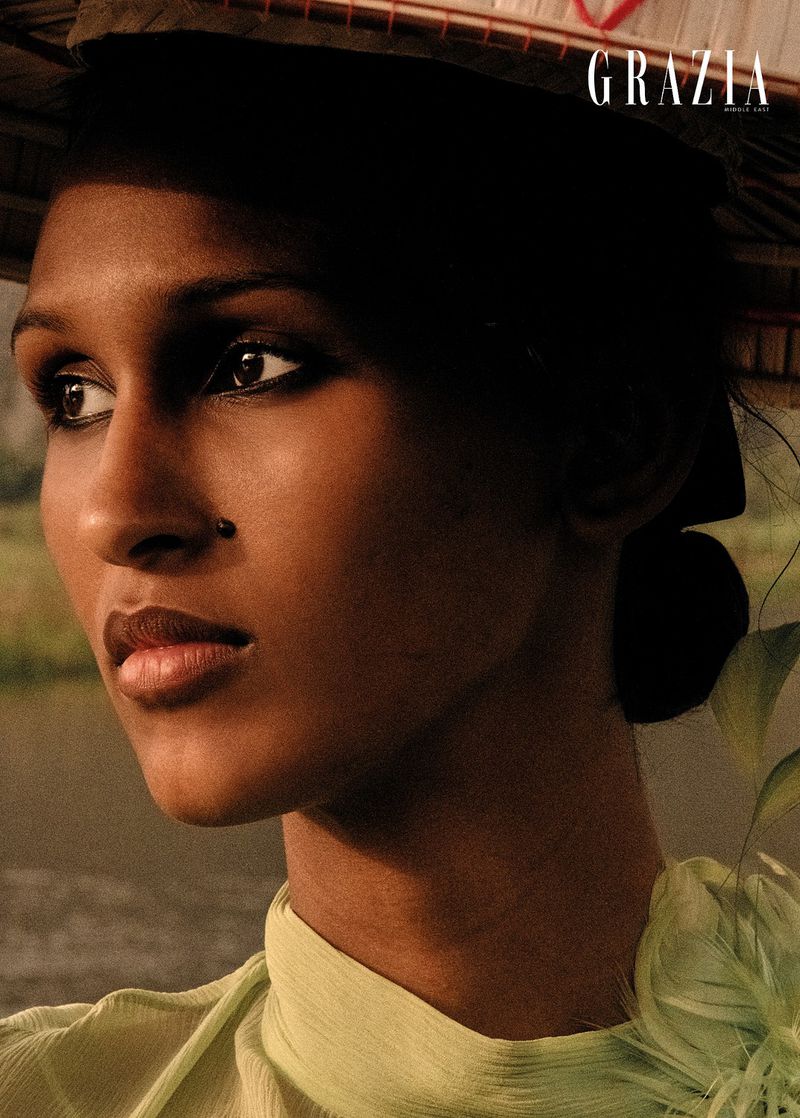Portrait of an adult woman with dark skin wearing a woven hat and light green top; she's looking to the side against a blurred outdoor background.