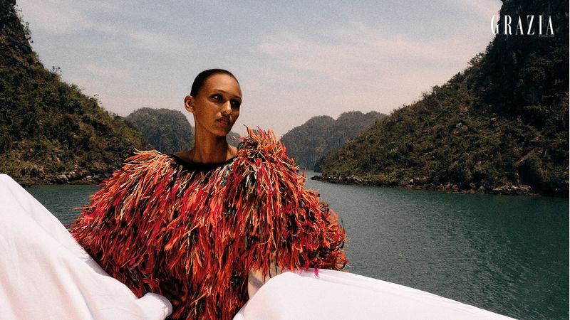 Fashion editorial shot of a woman in a fringed garment in a natural setting with mountains and a body of water as the backdrop.