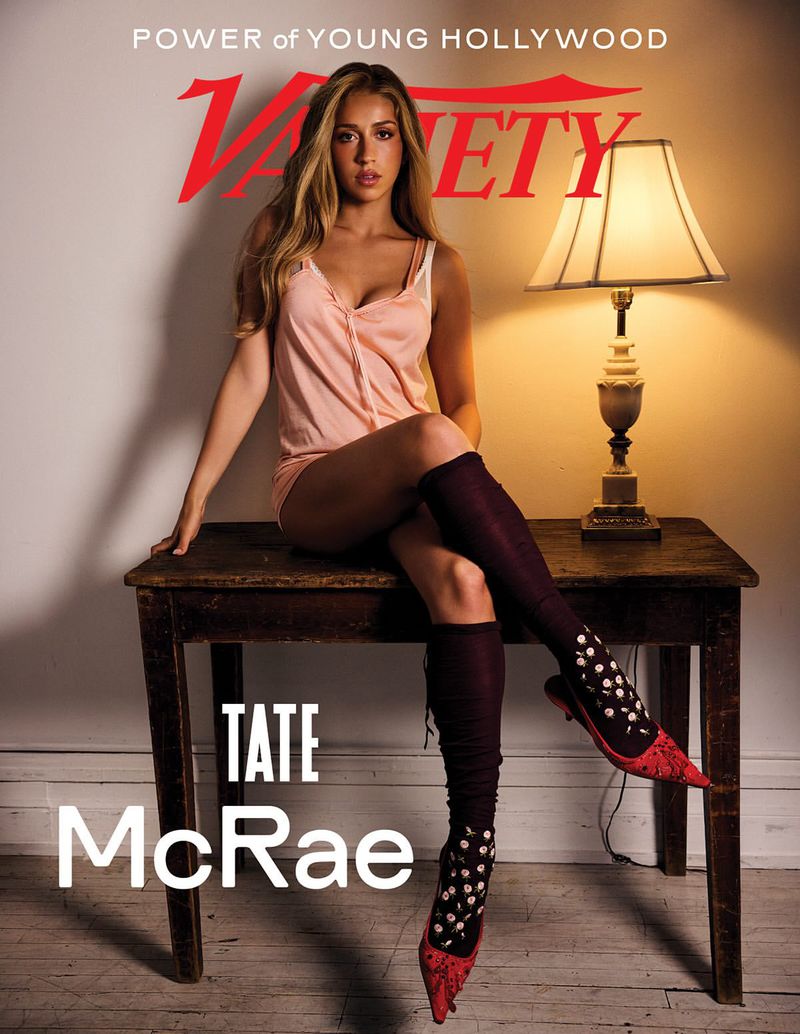 Tate McRae sits on a rustic wooden table wearing a pale pink tank top and knee-high dark socks with red pointed-toe shoes.