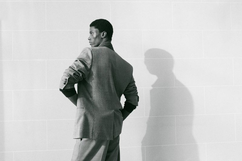 Black and white photo of a man in a tailored grey suit jacket facing away against a pale block wall casting a sharp shadow.