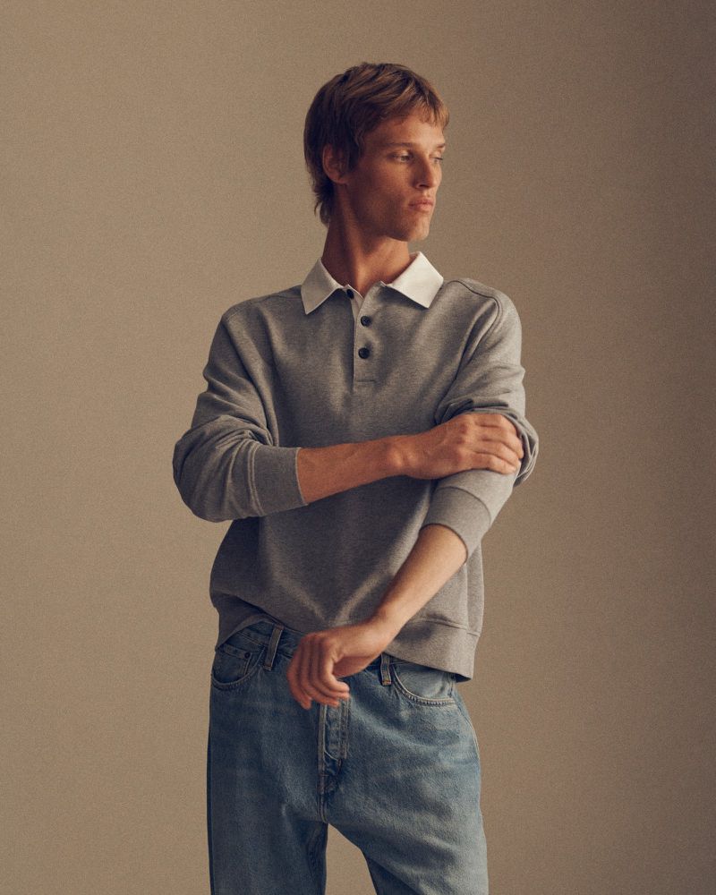 Model wearing a grey long-sleeve polo sweatshirt with a white collar and blue jeans against a neutral studio background.