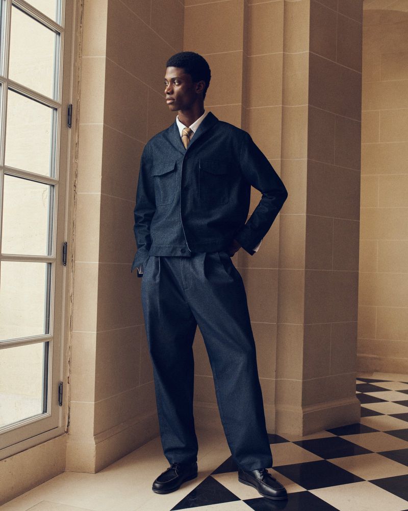 A Black man standing by a tall window wearing a navy blue tailored jacket, matching pleated trousers, and a tie over a white shirt.