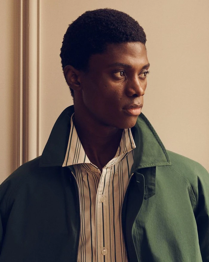 Black man wearing a dark green collared jacket over a striped button-down shirt against a warm beige background.
