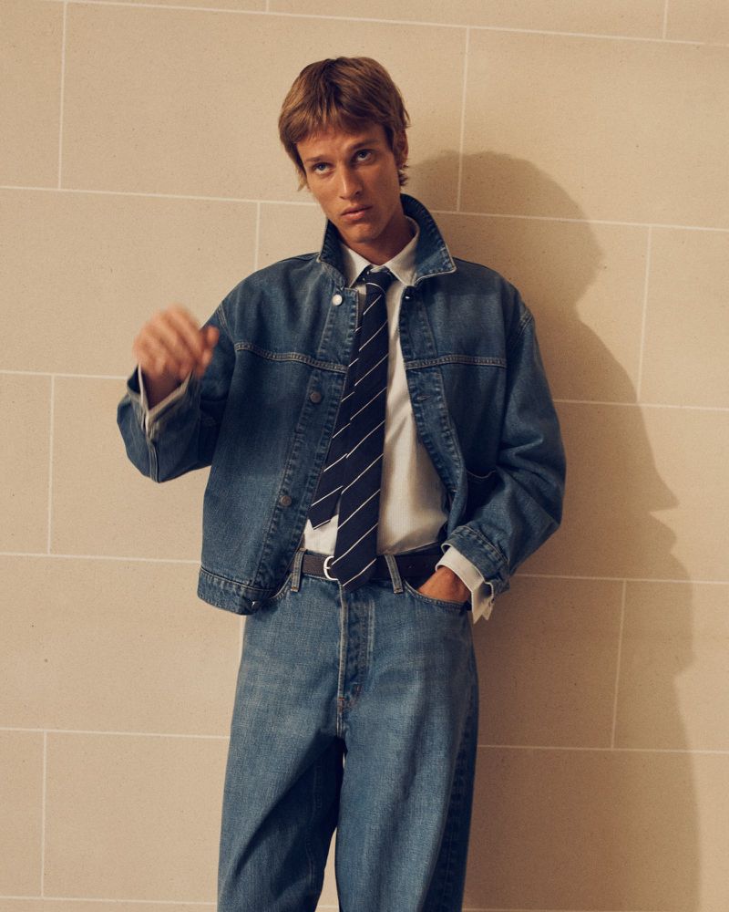 Man wearing a blue denim jacket, matching jeans, white collared shirt, and striped tie against a light stone block wall.
