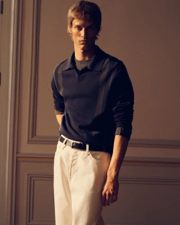 A man wearing a navy collared polo sweater and white trousers posing against a neutral wall with molding.
