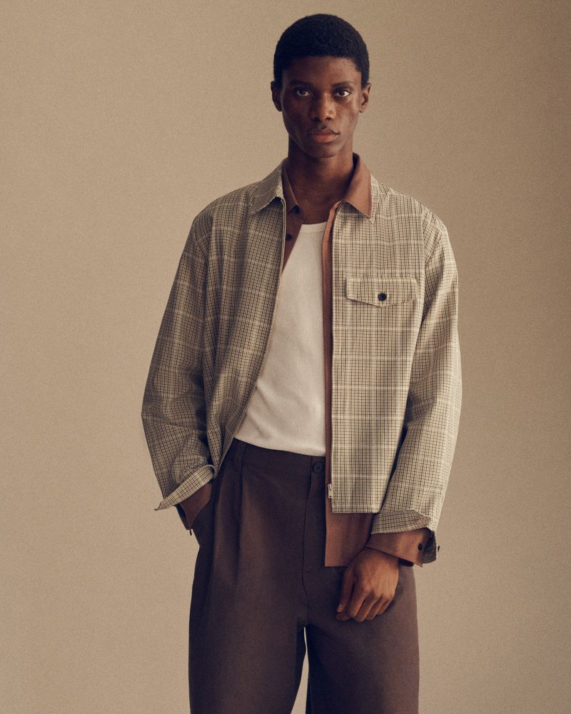 Black man wearing a beige plaid zip-up overshirt over a brown shirt, white top, and pleated brown trousers against a warm backdrop.