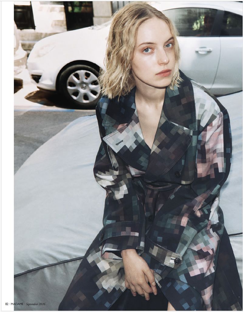 Model Sylwia Kuta seated wearing a pixelated patterned trench coat outdoors near a parked white car.