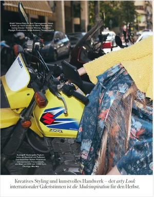 Model Sylwia Kuta in a textured yellow top and fringed silk skirt leaning on a yellow Suzuki motorcycle on an urban street.