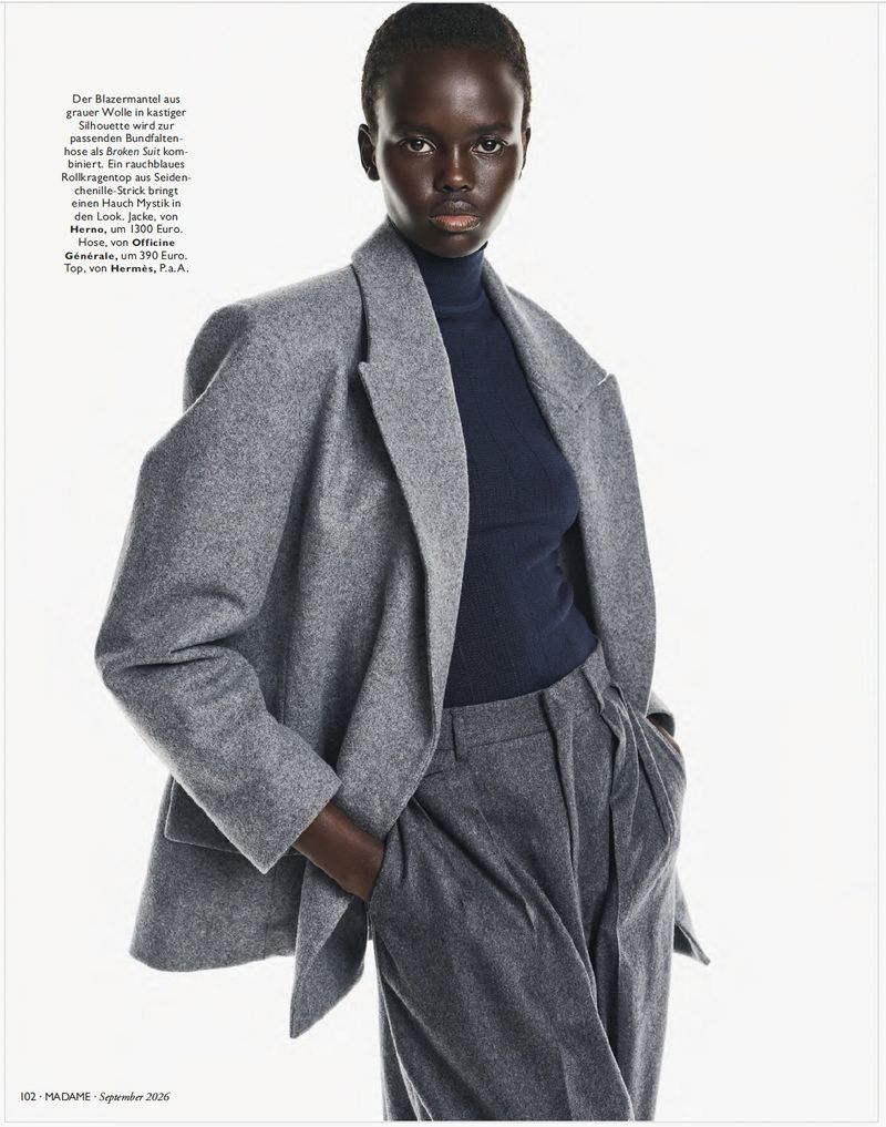 Model Ajak Dhieu wearing an oversized grey wool blazer, navy turtleneck, and pleated trousers against a white background.