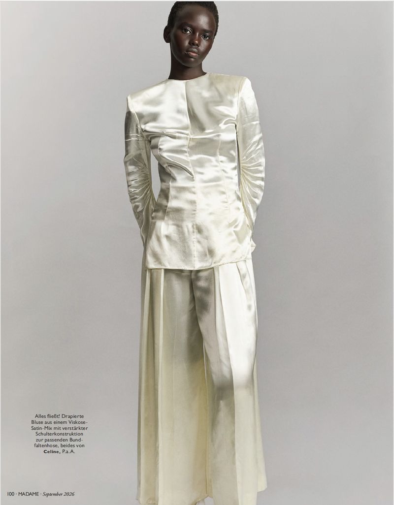 A Black model stands against a plain grey backdrop wearing a lustrous cream satin long-sleeved top and wide-leg trousers by Celine.