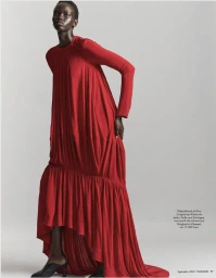 Model Ajak Dhieu wearing a flowing long-sleeved red pleated gown and black kitten heels against a light grey studio background.