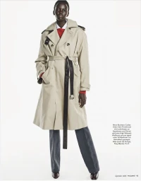 Model Ajak Dhieu wearing a belted beige trench coat layered over grey trousers against a solid white background.