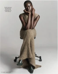 Model Ajak Dhieu seated on a stool wearing a sleeveless taupe knit maxi dress against a light grey studio background.