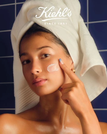 Close-up of a woman with fair skin, wearing a towel on her head, applying cream to her cheek. The Kiehl's logo is visible in the background.