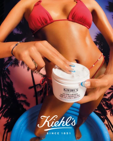 Woman in red bikini applies Kiehl's Ultra Body Mega Moisture Squalane Cream in an indoor setting with a palm tree background.