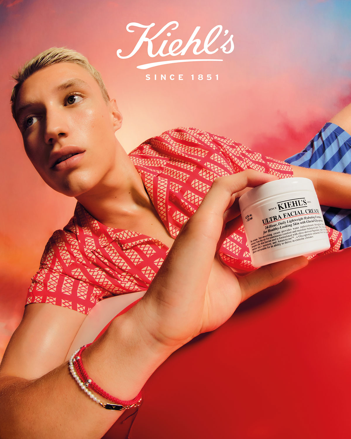 A fair-skinned young adult male poses with Kiehl's Ultra Facial Cream in a beauty advertisement with a pink, orange, and white gradient background.
