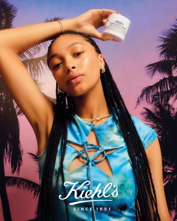A woman poses with Kiehl's skincare product against a backdrop of palm trees silhouetted against a purple and orange sky.