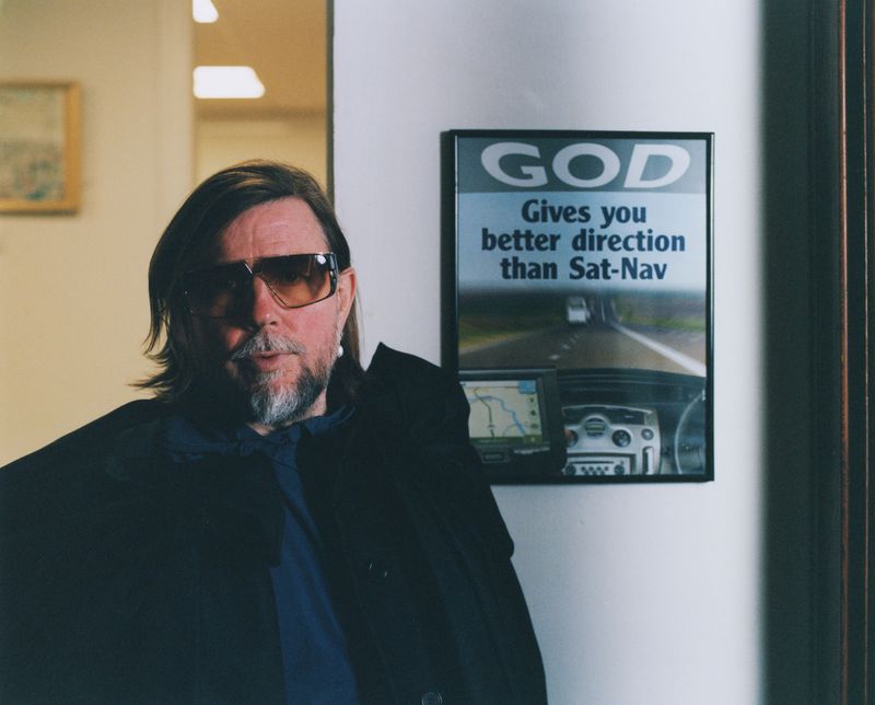 Portrait of an adult male wearing sunglasses and a dark jacket, posed next to a framed advertisement in an indoor environment.