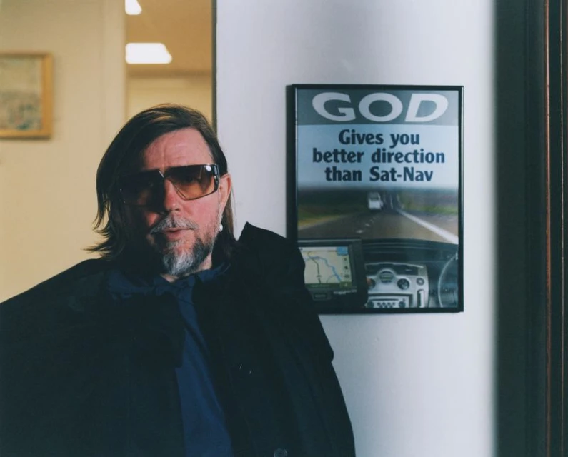 Portrait of an adult male wearing sunglasses and a dark jacket, posed next to a framed advertisement in an indoor environment.