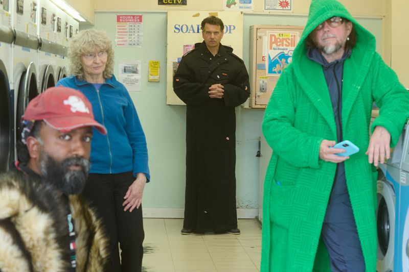Four people stand in a laundromat. One wears a red hat and fur coat, another a blue jacket, another a black robe, and another a green robe.