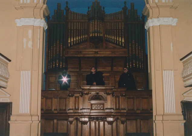 Three men in formal wear standing behind a large wooden organ in an ornate interior, possibly a church or concert hall. 