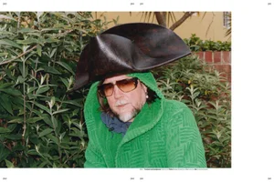 Man wearing a green textured hooded robe, large dark hat, and sunglasses standing outdoors against leafy bushes