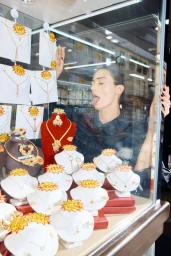 Model Maeva Marshall presses her tongue against a jewellery store display window surrounded by gold necklaces on bust stands.