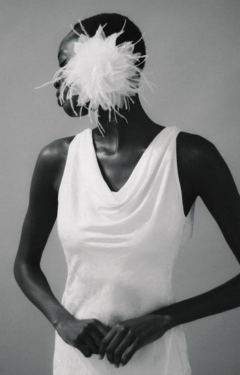 A Black woman with short hair wearing a white cowl neck dress and a large, white feathered floral hairpiece covering part of her face.