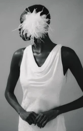 Black woman in a white cowl-neck dress wearing a large white feather headpiece over her profile against a studio backdrop.